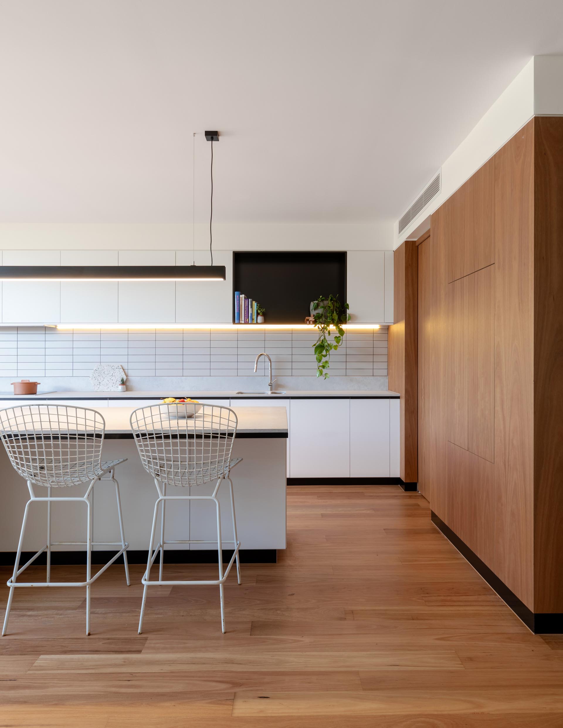 Современная кухня включает минималистские белые шкафы, черную акцентную полку, большой остров с местом для сидения и нижнее освещение.