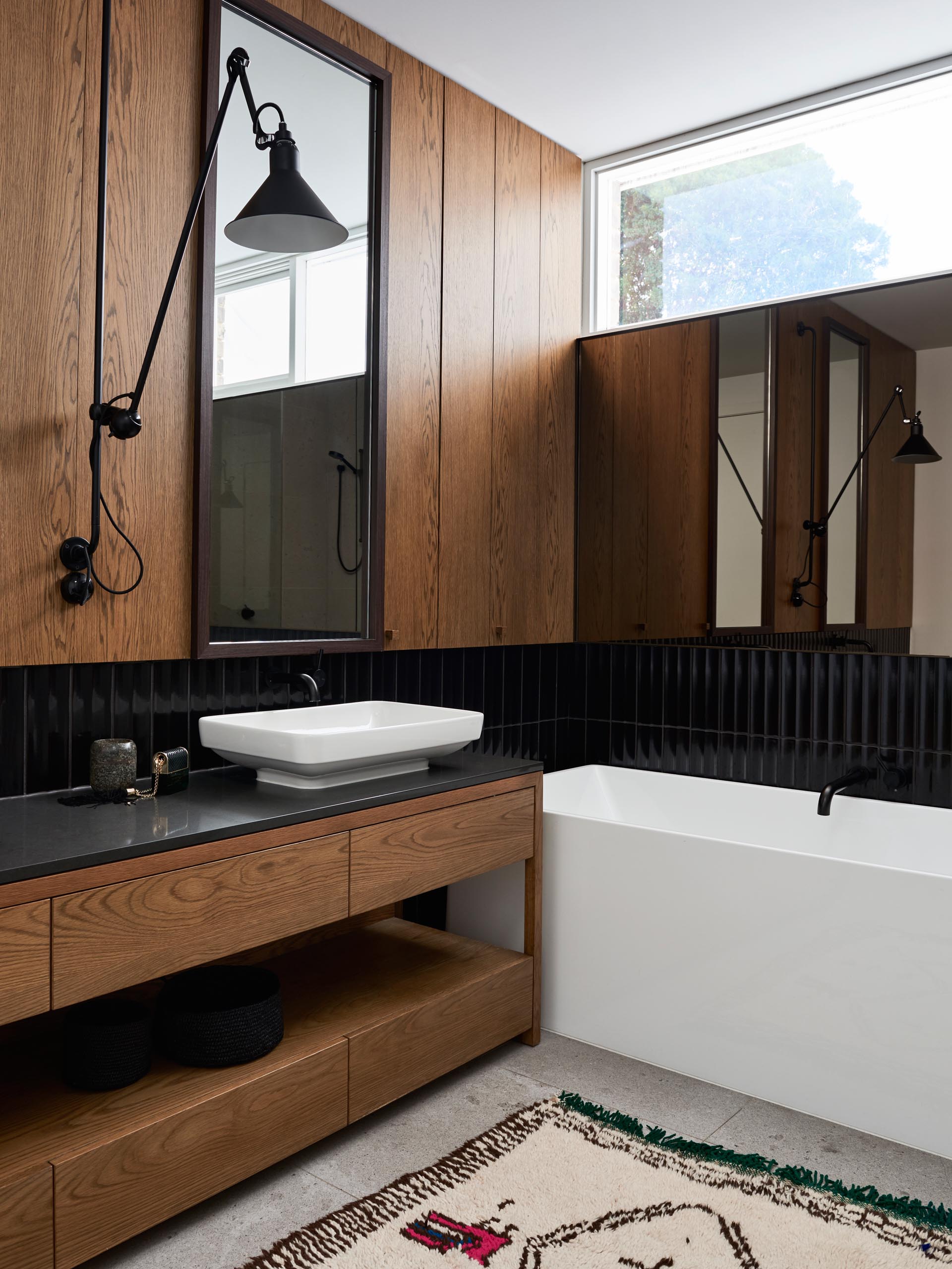 В этой современной ванной комнате деревянные панели и деревянный туалетный столик сочетаются с черной плиткой, белым умывальником и белой ванной.