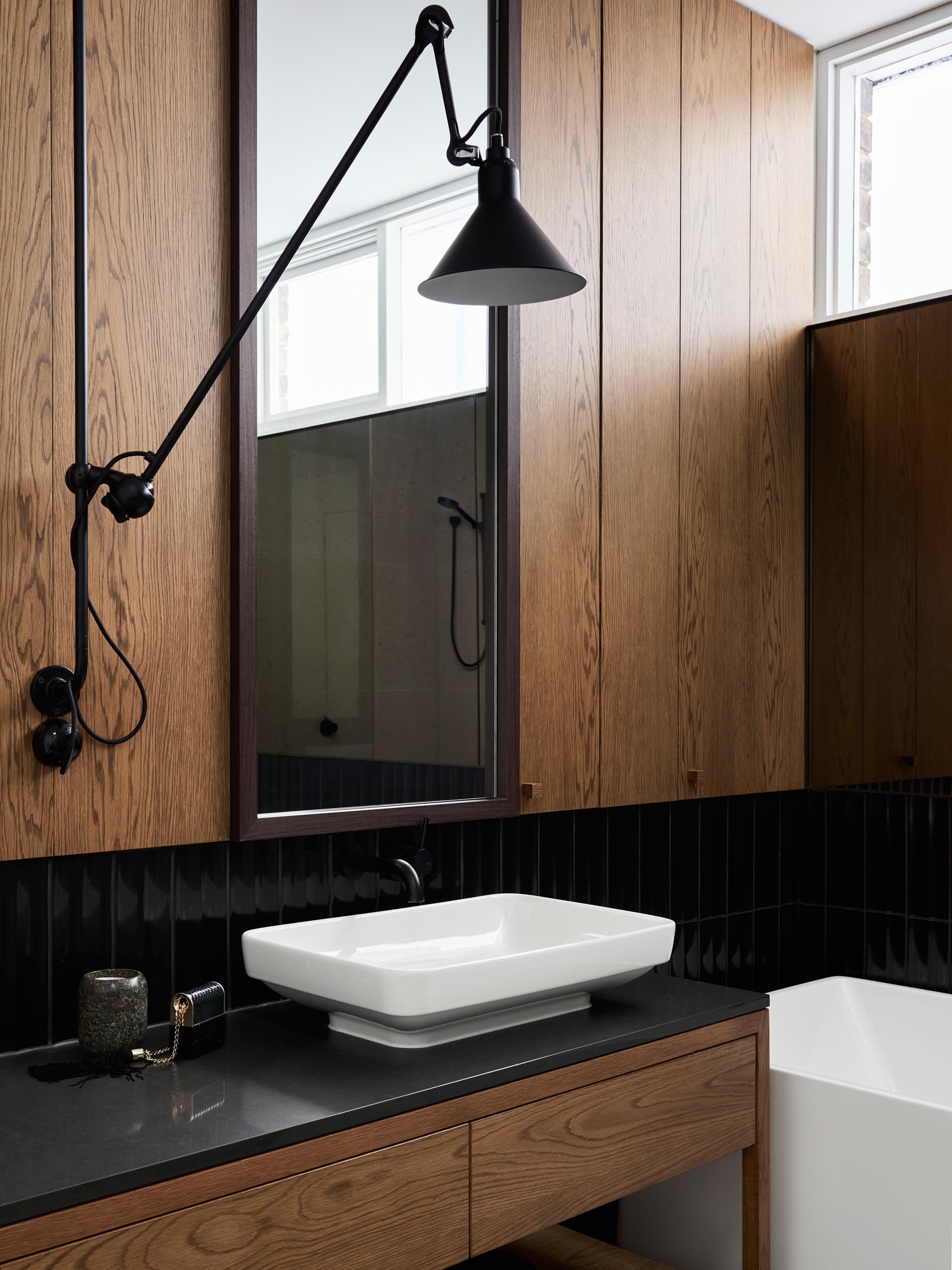 В этой современной ванной комнате деревянные панели и деревянный туалетный столик сочетаются с черной плиткой, белым умывальником и белой ванной.