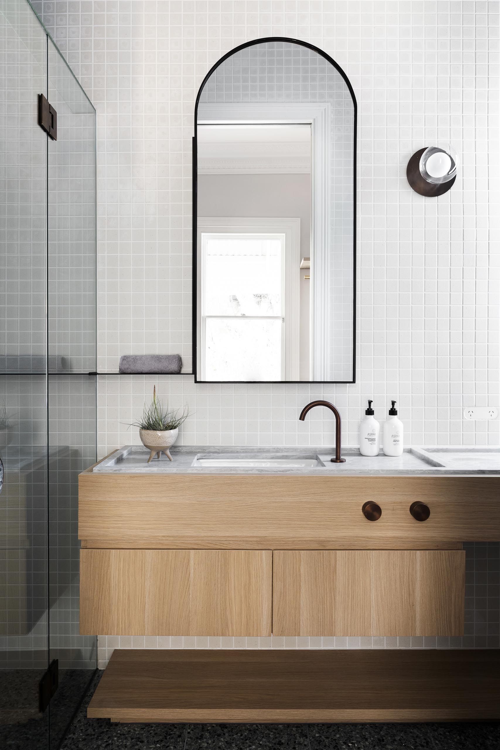 Современная ванная комната с деревянным туалетным столиком, кафельной стеной и арочным зеркалом.