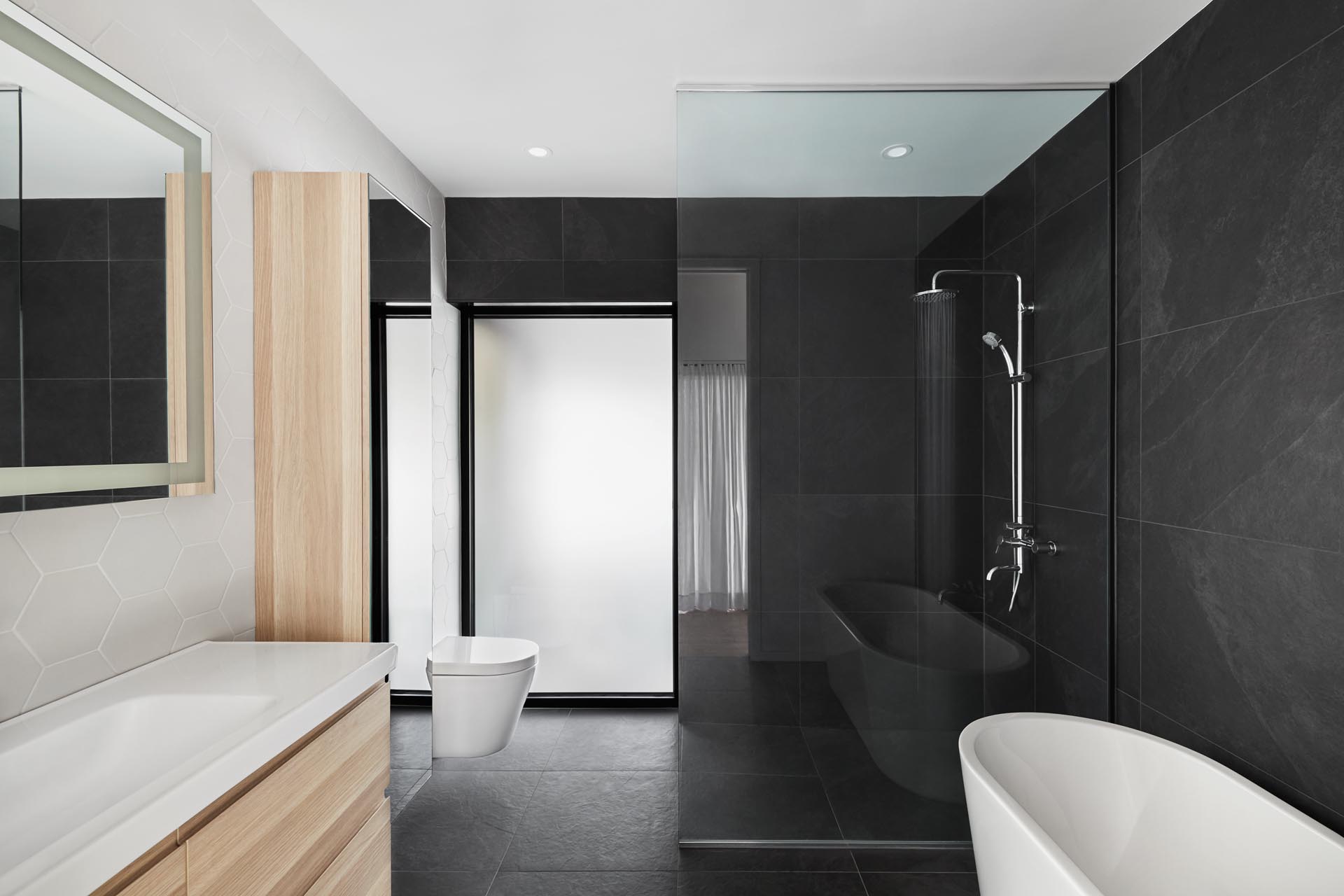 В этой современной ванной комнате широкоформатная черная плитка покрывает две стены, в то время как есть туалетный столик из светлого дерева и стена из белой шестиугольной плитки.