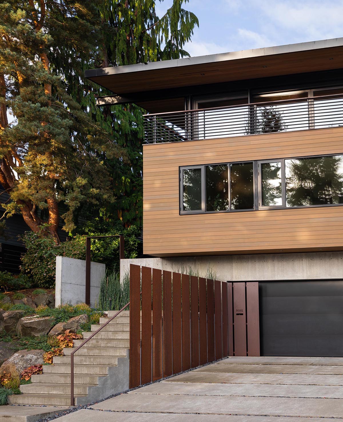 Фасад этого современного дома украшен бетоном, черной гаражной дверью, стальными элементами, деревянным сайдингом и ступенями, ведущими к входной двери.