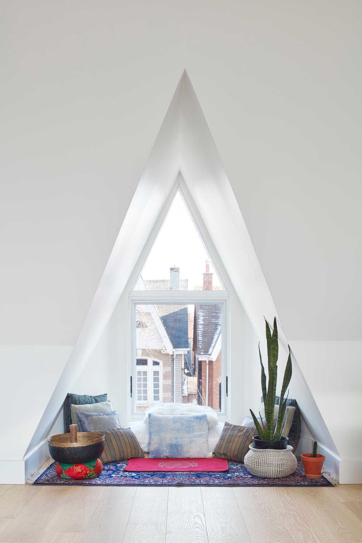 Уголок у окна треугольной формы, обставленный ковриком, множеством подушек, растений и чашей для медитации.