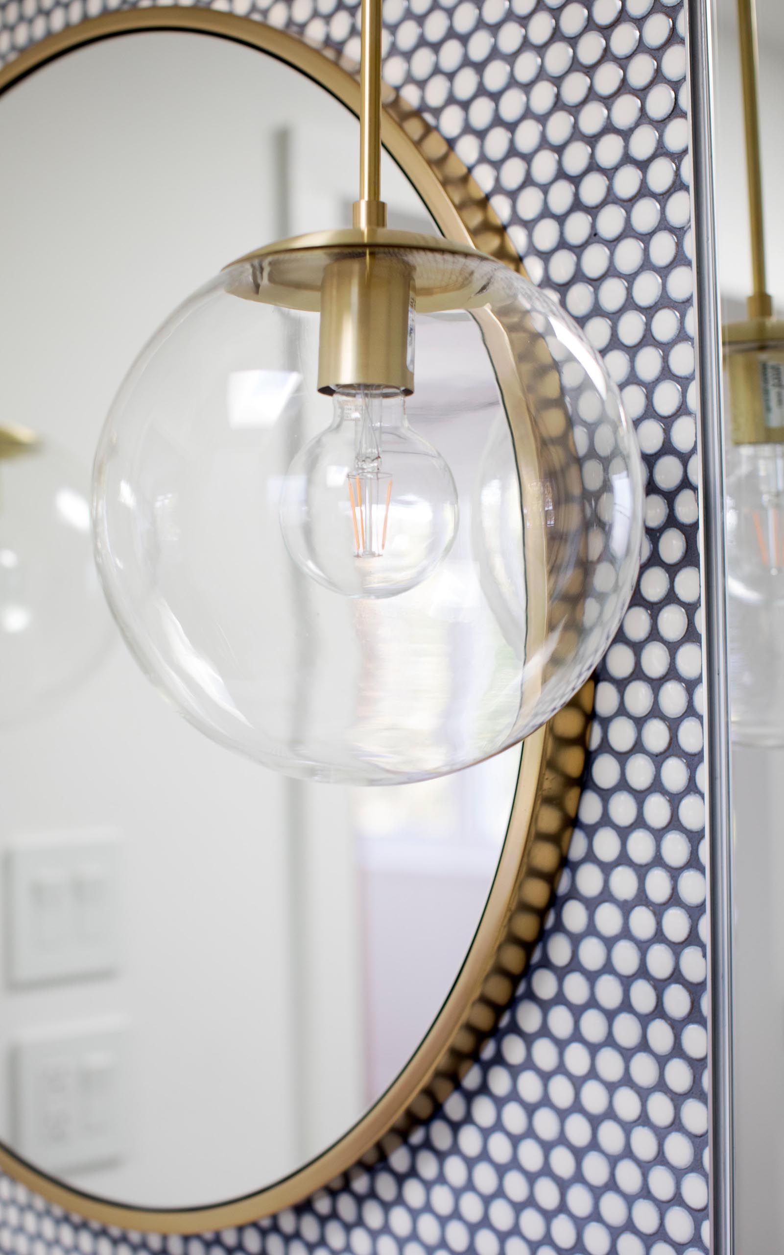 Современная ванная комната с белой плиткой для пенни, контрастной темной затиркой, круглым зеркалом в золотой раме и подходящим подвесным светильником со стеклянным шаром.