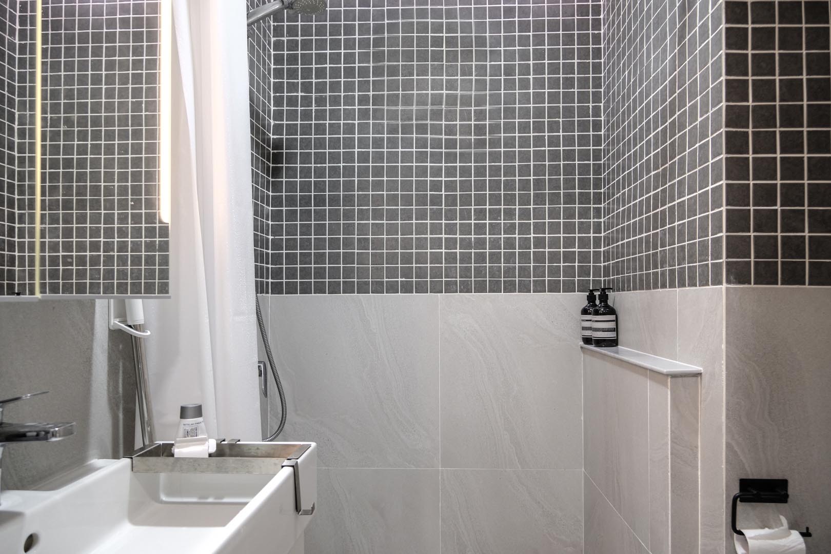 В этой маленькой ванной комнате большая плитка покрывает нижнюю половину стен, а верхняя половина покрыта квадратной серой плиткой.