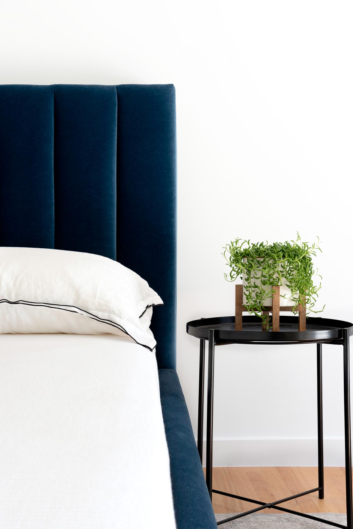 Спальня с глубоким королевским синим бархатным изголовьем и каркасом кровати, а также матовым черным прикроватным столиком с небольшим растением.
