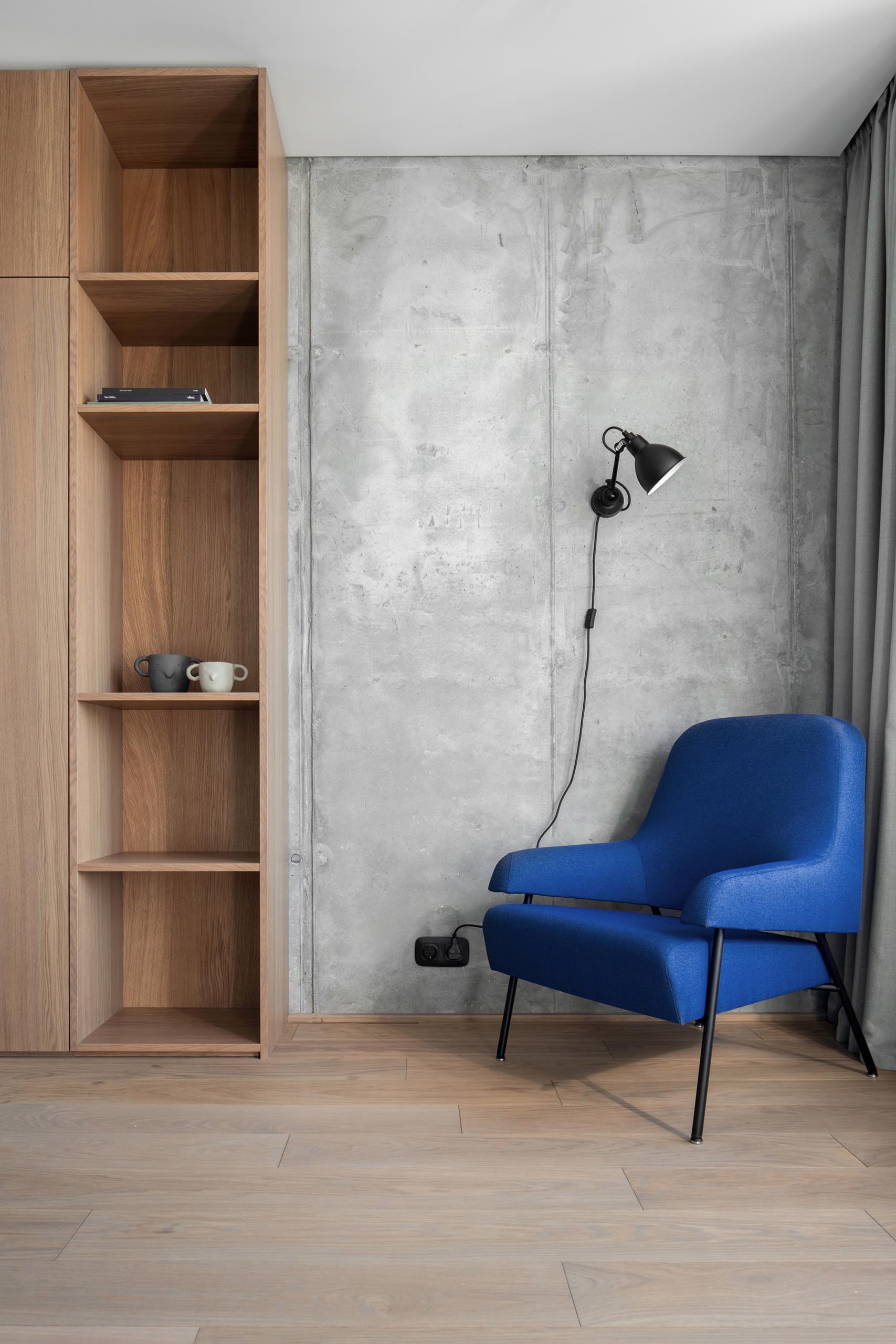 Современный домашний офис с длинными шторами, синим креслом, деревянными стеллажами и бетонной стеной.